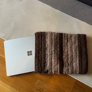 Könnte beinhalten: Eine braune gestrickte Laptop-Hülle mit einem dezenten Streifenmuster. Die Hülle liegt auf einer Holzoberfläche, auf der ein Laptop teilweise sichtbar ist.