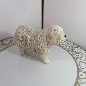 Op de afbeelding: Een wit keramiek beeldje van een Komondor-hond. De hond staat op een wit bord met een gouden rand.