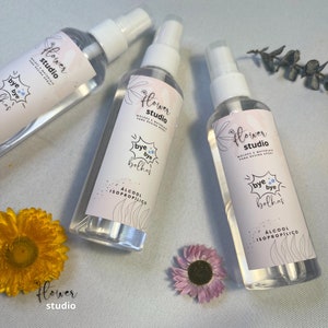 May include: Three clear plastic spray bottles with white labels. The labels have the text "Flower Studio" and "Bye Bye Bolhas" with a blue and white graphic of a bubble. The bottles are labelled "Alcohol Isopropilico".