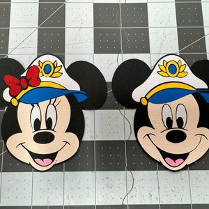 May include: Two paper cutouts of Mickey Mouse and Minnie Mouse wearing blue and white sailor hats with gold accents. The hats have a yellow band and a gold emblem with a yellow star.