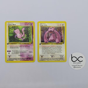 May include: Two Pokémon trading cards featuring "Failed Mew" and "Pixel Ditto" characters. The cards have yellow borders and include character names, HP, and special abilities. A white sticker with "BCD CARDS" and social media information is also visible.