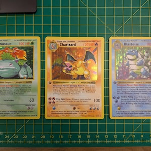 Puede incluir: Tres cartas coleccionables Pokémon vintage: Venusaur, Charizard y Blastoise. Las cartas tienen un acabado holográfico y presentan ilustraciones coloridas de los personajes Pokémon. Las cartas se muestran en una esterilla de corte verde.