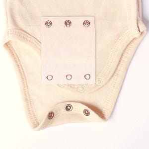 Puede incluir: Body de bebé color crema con cierres de corchete. El body presenta un parche de tela rectangular con tres corchetes en la parte superior y tres en la inferior. Corchetes adicionales ubicados en los hombros y la entrepierna.