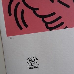 May include: A pink and green print with black outlines of a figure with a raised arm. The print is numbered 3/100 and signed "Keith Haring" with a sunburst logo.