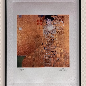 May include: A framed print of a painting by Gustav Klimt, featuring a woman in a gold dress with a geometric pattern. The painting is titled "The Lady in Gold" and is numbered 106/300.