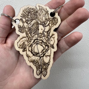 May include: A wooden keychain featuring a laser-engraved image of two anime characters, one with spiky hair and the other with a long, flowing mane, both in mid-air with their fists raised.