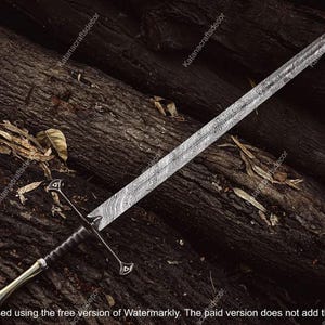 May include: A long, silver and black sword with a detailed blade pattern rests on dark brown wooden logs. The sword features a crossguard and a decorative handle. The blade has a textured, patterned design.