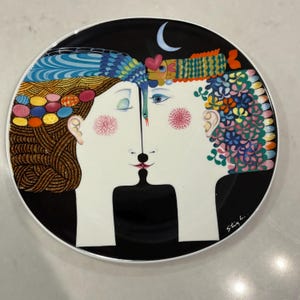 Op de afbeelding: Decoratieve keramische plaat met een zwarte achtergrond en een witte rand. De plaat toont een gestileerde illustratie van twee gezichten, met kleurrijk haar en bloemaccenten. Een halve maan en een hart zijn ook afgebeeld.