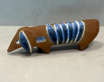 Puede incluir: Figura de perro salchicha de cerámica en tonos marrón y azul. El perro tiene un cuerpo largo con detalles de rayas azules y blancas en la espalda. La figura tiene un ojo pequeño y redondo y una etiqueta que dice "DESIGN LISA LARSON."