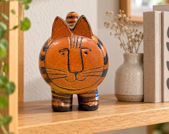 May include: A ceramic cat figurine with a round orange body and black stripes. The cat has pointed ears, a simple hand-drawn face with eyes, a nose, and whiskers. A small blue rectangular label is visible on the lower right leg of the cat.