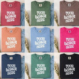 May include: Nine colorful t-shirts in various shades, including red, green, brown, orange, blue, pink, navy, teal, and black. Each shirt features the text "YOUR DESIGN HERE" in a white, stylized font. The shirts are folded and placed on top of denim jeans.