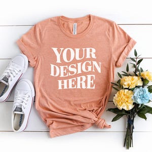 May include: A peach colored t-shirt with the text "YOUR DESIGN HERE" printed in white on the front. The shirt is laid flat on a white surface with a bouquet of yellow and blue flowers and a pair of white sneakers.