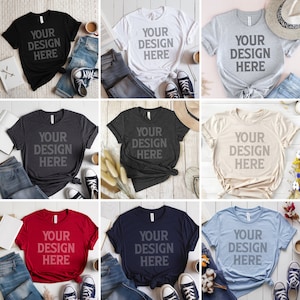 May include: Nine different colour t-shirts with the text "YOUR DESIGN HERE" printed on the front. The t-shirts are laid out on a white wooden surface with various props, including jeans, hats, and flowers.