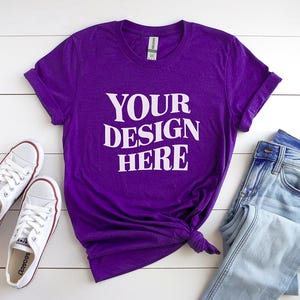 以下が含まれることがあります： 白い文字で「YOUR DESIGN HERE」と書かれた紫色のTシャツ、水色のジーンズ、白いスニーカーの組み合わせ。Tシャツは裾で結ばれています。スニーカーには赤いアクセントがあります。