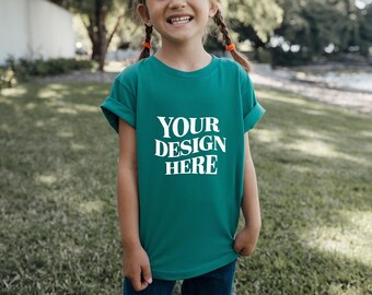 以下が含まれることがあります： 白い「YOUR DESIGN HERE」の文字が入った、袖をまくったターコイズ色のTシャツ。芝生の上に立っている人が着ています。背景には木と水面があります。
