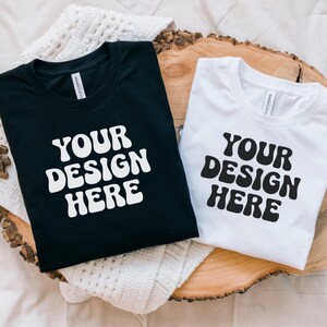 Könnte beinhalten: Zwei T-Shirts, eines schwarz und eines weiß, mit dem Text "YOUR DESIGN HERE" in einer Retro-Schriftart. Die Shirts sind gefaltet und auf einer Holzoberfläche ausgelegt.