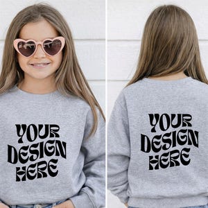 Peut inclure: Un sweat-shirt gris clair avec le texte "YOUR DESIGN HERE" en noir, présenté de face et de dos. La vue de face montre une personne portant des lunettes de soleil roses en forme de cœur. Le sweat-shirt est de style col rond.