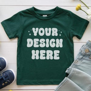 May include: A dark green t-shirt with short sleeves and a white design that says "YOUR DESIGN HERE" on a white wooden background.