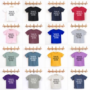 Pode incluir: Uma coleção de camisetas de várias cores, cada uma exibindo o texto "YOUR DESIGN HERE" em uma cor contrastante. As camisas são dispostas em cabides de madeira contra um fundo branco. As cores incluem branco, preto, azul marinho, vermelho, rosa, bordô, azul, cinza, verde, verde-azulado, amarelo, roxo e cinza claro.