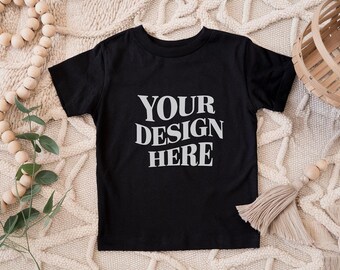 Könnte beinhalten: Schwarzes T-Shirt mit dem Text "YOUR DESIGN HERE" in weißer, stilisierter Schrift. Das Shirt wird auf einer strukturierten, cremefarbenen Oberfläche präsentiert, mit dekorativen Holzperlen und einem geflochtenen Korb im Hintergrund. Ein farblich neutraler Quastenanhänger ist ebenfalls sichtbar.