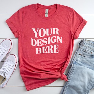 May include: Red t-shirt with the text "YOUR DESIGN HERE" in white, paired with light blue jeans and white sneakers. The t-shirt is knotted at the waist, and the sneakers have red accents.