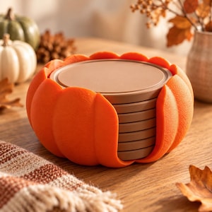 May include: A stack of round, brown coasters housed within a bright orange, pumpkin-shaped holder. The holder features a textured, ribbed exterior that mimics the appearance of a pumpkin, while the coasters are smooth, flat, and circular.