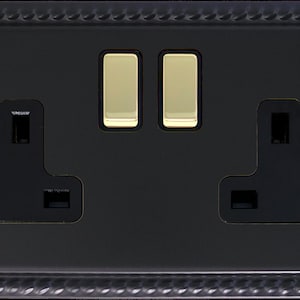 May include: A double electrical wall socket with a matte black finish and a rope-style border. The unit features two black three-pin sockets and two polished brass rocker switches positioned in the centre, secured by two matching black screws.