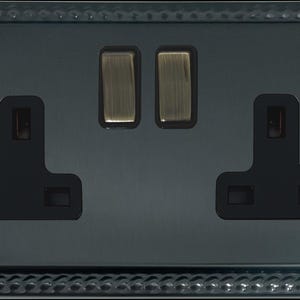 May include: A double electrical wall socket with a dark metallic finish and a decorative beaded border. The unit features two black socket inserts and two vertical metallic switches positioned in the center, secured by two flat-head screws on the sides.
