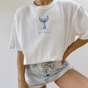 May include: White t-shirt with a blue whale tail graphic and the text "ALL IS WHALE" printed on the front.