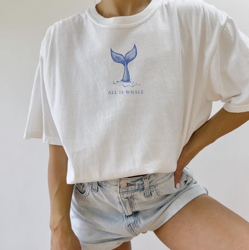 May include: White t-shirt with a blue whale tail graphic and the text "ALL IS WHALE" printed on the front.