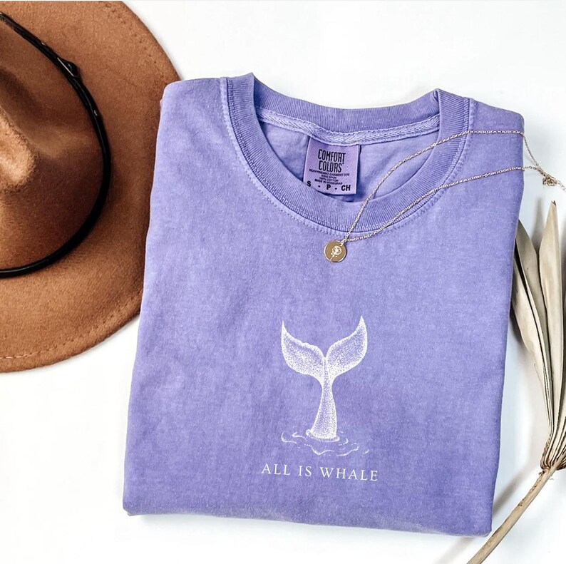 May include: A lavender t-shirt with a white whale tail graphic and the text "All is Whale".