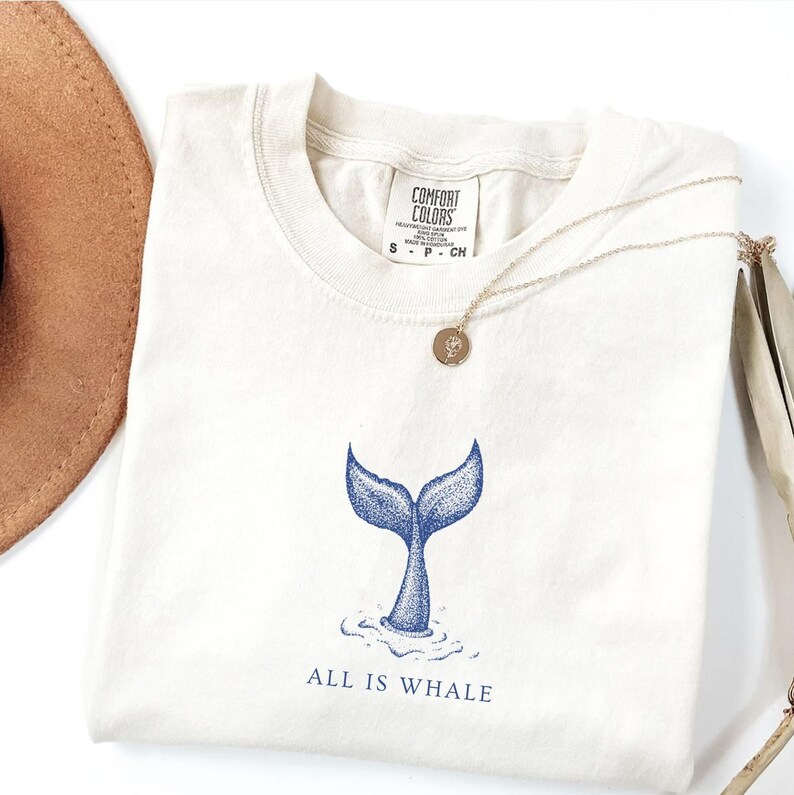 May include: A white t-shirt with a blue whale tail graphic and the text "ALL IS WHALE".