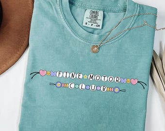 May include: A teal Comfort Colors t-shirt with the words "FINE MOTOR CLUB" in a beaded design. The design includes hearts, flowers, and smiley faces. A gold necklace with a pendant is draped across the shirt.