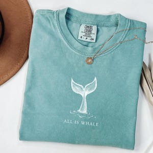 May include: A teal blue t-shirt with a white whale tail graphic and the text "ALL IS WHALE".