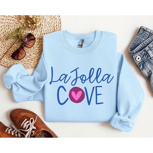 May include: A light blue sweatshirt with the text "La Jolla Cove" in blue lettering and a pink heart in the middle of the word "Cove".