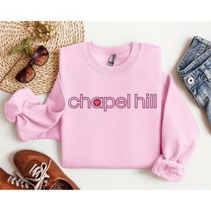 May include: Pink sweatshirt with a maroon graphic of the text "chapel hill" with a small red heart in the middle of the word "chapel".