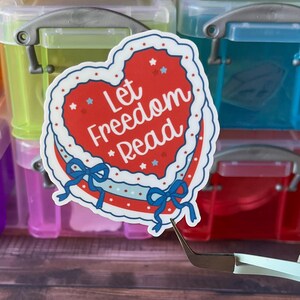 May include: A heart-shaped sticker with the text "Let Freedom Read" in red, white, and blue. The sticker has a decorative border and blue bows. Several stickers are visible, along with colorful plastic storage boxes.