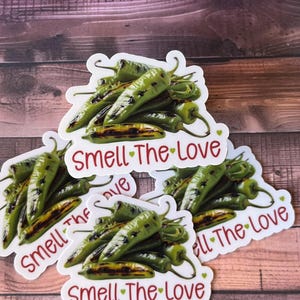 May include: White stickers featuring a cluster of green chili peppers with charred tips. The text "Smell The Love" is written in red script on each sticker. The stickers are on a wooden surface.