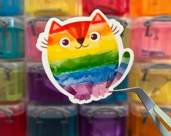 May include: A colorful sticker of a cat with a rainbow design. The cat has a heart-shaped nose and is sitting. The sticker is being held by a pair of tweezers. The background is a collection of small, colorful containers.