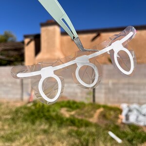 May include: A clear, transparent sheet with three white-outlined circles connected by white lines, resembling eyeglasses. A pair of tweezers holds the sheet against a blurred outdoor background.