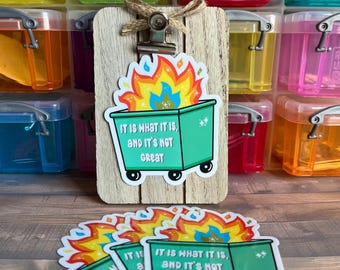 May include: Four green and white garbage can stickers with flames and the text "IT IS WHAT IT IS, AND IT'S NOT GREAT". The stickers are on a wooden surface.