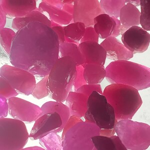 Könnte beinhalten: Eine Sammlung von rosafarbenen Rubingranaten, einige mit einem durchscheinenderen Aussehen als andere. Die Edelsteine ​​haben verschiedene Formen und Größen und sind auf einer weißen Oberfläche verstreut.