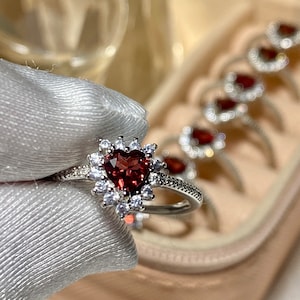 Op de afbeelding: Een zilveren ring met een hartvormige rode edelsteen omringd door kleinere heldere edelstenen. De ring wordt gepresenteerd in een sieradendoos.