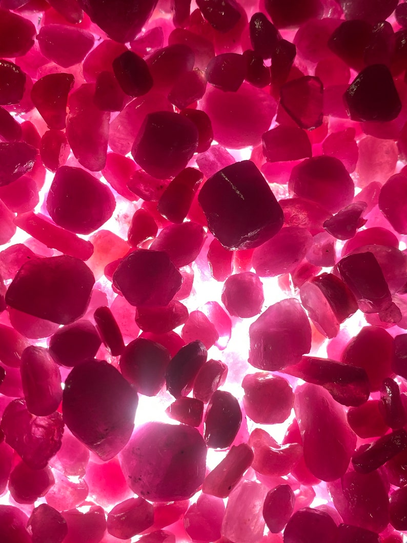 Puede incluir: Una vista cercana de una colecci&oacute;n de piedras preciosas rosadas y rojas. Las piedras preciosas son de varias formas y tama&ntilde;os, y est&aacute;n esparcidas sobre una superficie blanca.