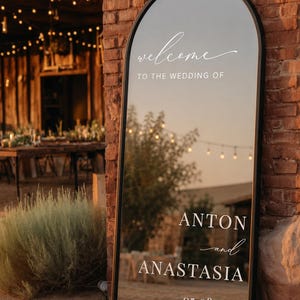 May include: A tall, arched floor mirror with a black frame featuring white lettering that reads: welcome TO THE WEDDING OF ANTON and ANASTASIA 05.08.2027. The reflective surface displays a clear view of the surrounding outdoor environment.