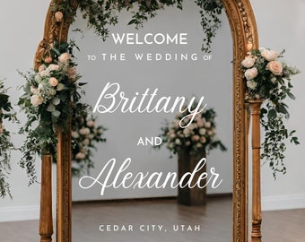 Puede incluir: Un espejo con marco dorado y decoraciones florales a cada lado. El espejo refleja un cartel que dice "Bienvenido a la boda de Brittany y Alexander, Cedar City, Utah".