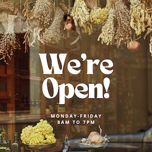 May include: A storefront window with a sign that reads "We're Open! Monday-Friday 8AM to 7PM". The window is decorated with dried herbs and flowers hanging from the top.