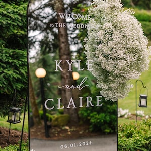 Op de afbeelding: Een zwarte spiegel met een witte booglijst, versierd met witte baby's breath bloemen. De spiegel toont de tekst "WELCOME to THE WEDDING KYLE and CLAIRE 06.01.2024".