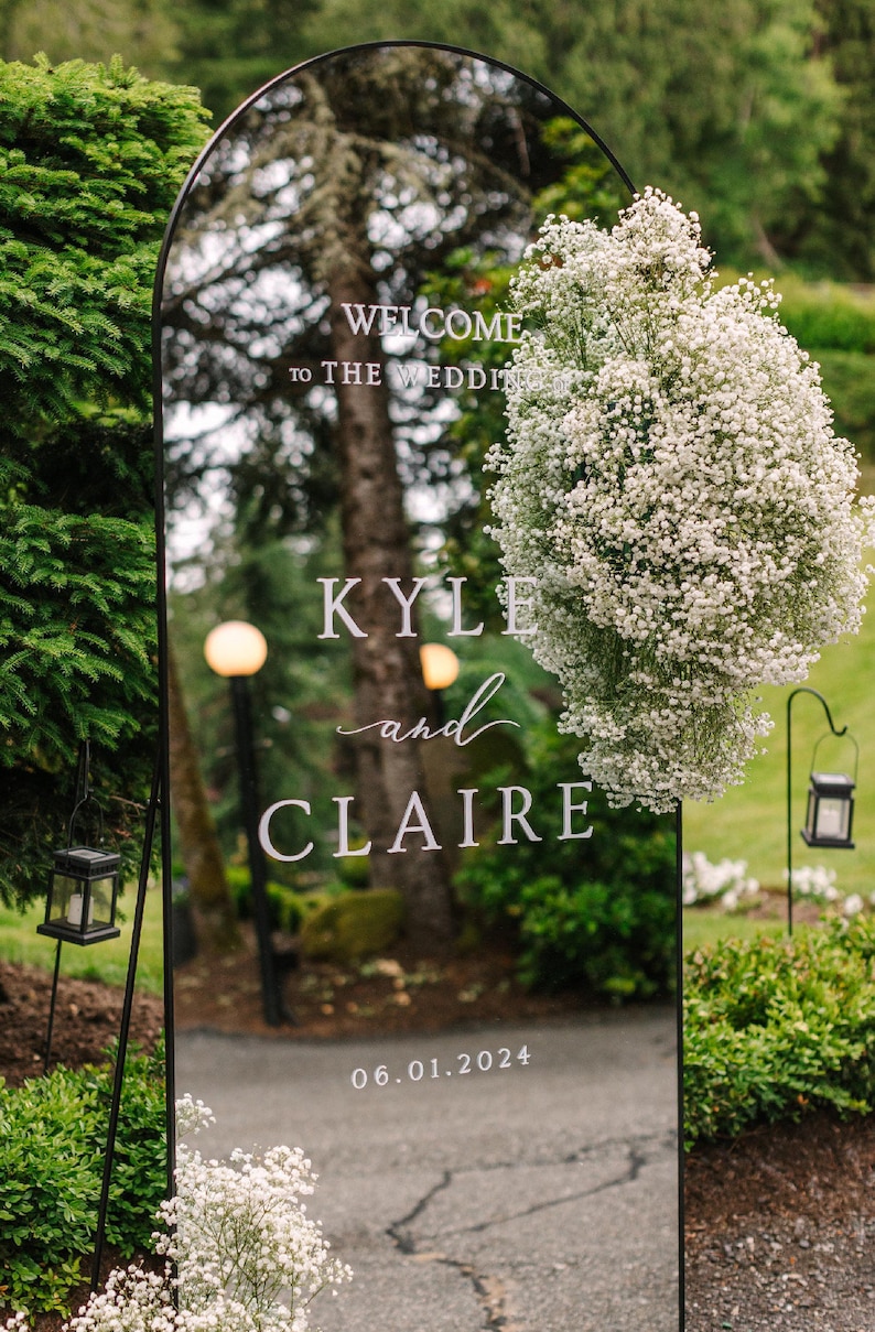 Op de afbeelding: Een zwarte spiegel met een witte booglijst, versierd met witte baby's breath bloemen. De spiegel toont de tekst "WELCOME to THE WEDDING KYLE and CLAIRE 06.01.2024".