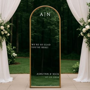 Puede incluir: Espejo dorado con forma de arco, con el texto "WE'RE SO GLAD YOU'RE HERE!" y los nombres "Ashlynn & Nick" con la fecha "07.25.2026". El espejo está situado sobre un fondo de vegetación y arreglos florales blancos.